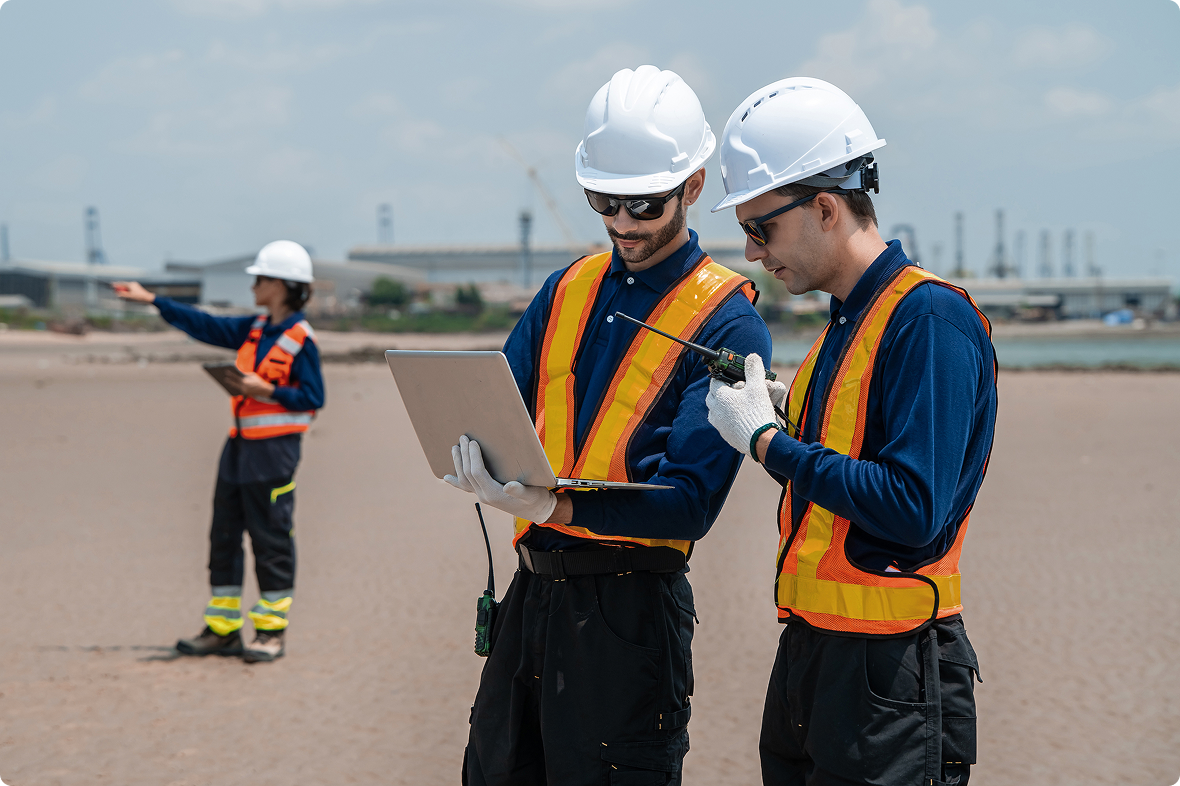 Two field technicians reviewing a laptop and radio at an industrial site, with a colleague gesturing in the background.