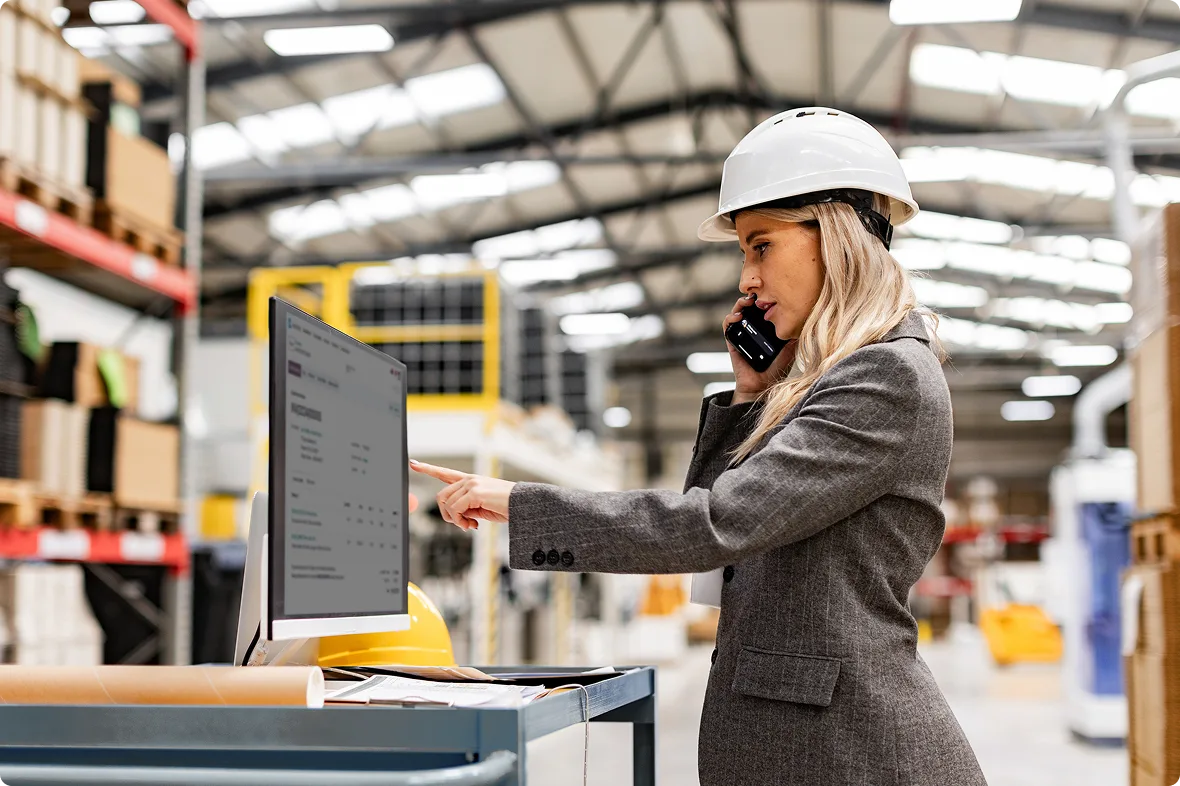 A woman in a hard hat on a phone call while pointing at equipment data on a monitor in a warehouse.