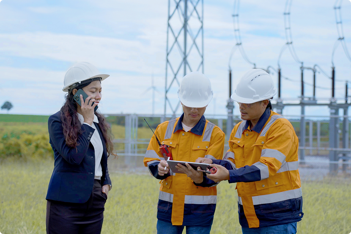 Two technicians in hi-vis jackets reviewing a tablet at an electrical substation while a colleague takes a phone call.